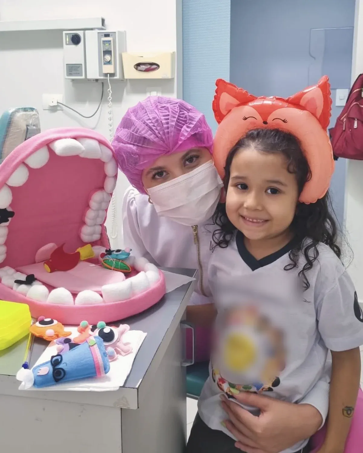 Uma menina com tiara de balão de raposa ao lado da boca gigante de feltro, com a dentista de touca rosa atrás dela.